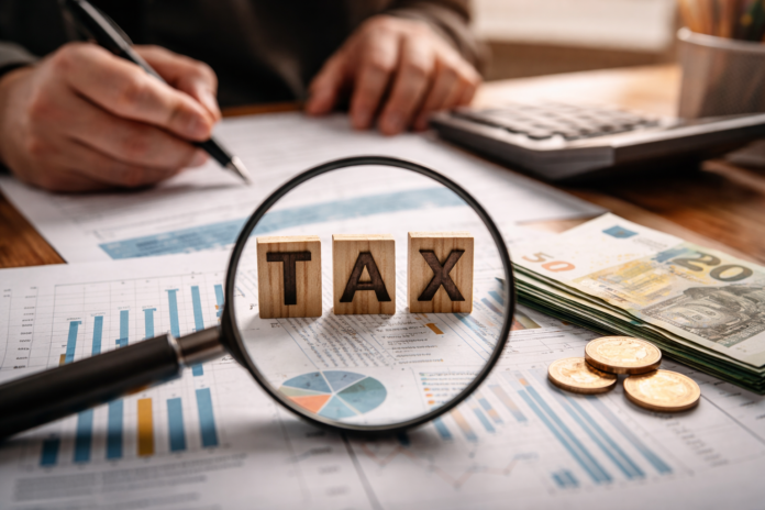 Μεγεθυντικός φακός εστιάζει στη λέξη TAX πάνω σε οικονομικά έγγραφα με ευρώ, αριθμομηχανή και γραφήματα, συμβολίζοντας τη φορολογική πολιτική και τον έλεγχο