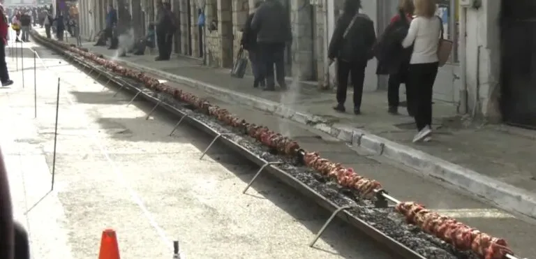 Μακριά υπαίθρια ψησταριά σε δρόμο με κοντοσούβλι πάνω από κάρβουνα, με κόσμο γύρω από τον χώρο.